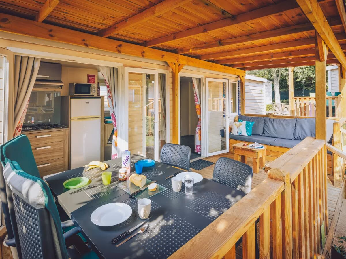 Gedekte eettafel op de overdekte veranda van een stacaravan, met de keuken en een loungebank erachter