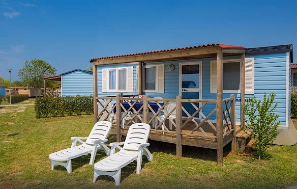 Lichtblauwe stacaravan met rood dak en een houten terras op Camping Park Umag, met twee witte ligbedden in het gras
