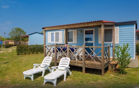 Lichtblauwe stacaravan met rood dak en een houten terras op Camping Park Umag, met twee witte ligbedden in het gras