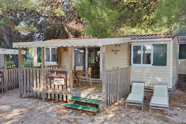 Oudere stacaravan met een naambordje SunLodge en een luifel boven het houten terras, tussen de dennen op Camping Park Umag