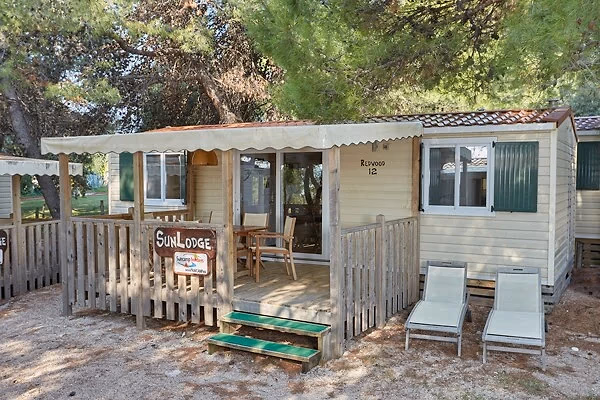 Oudere stacaravan met een naambordje SunLodge en een luifel boven het houten terras, tussen de dennen op Camping Park Umag