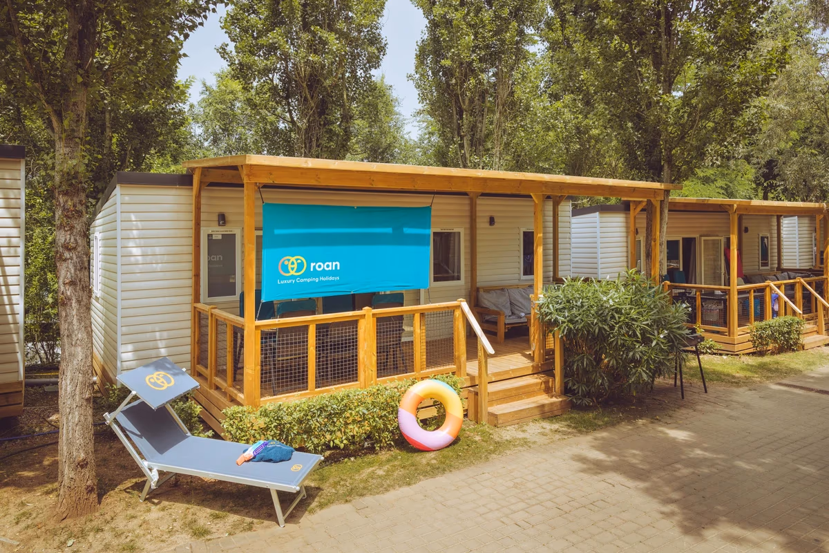 Stacaravan met een brede houten overkapping en een doek als zonwering, met een ligbed op het pad ervoor
