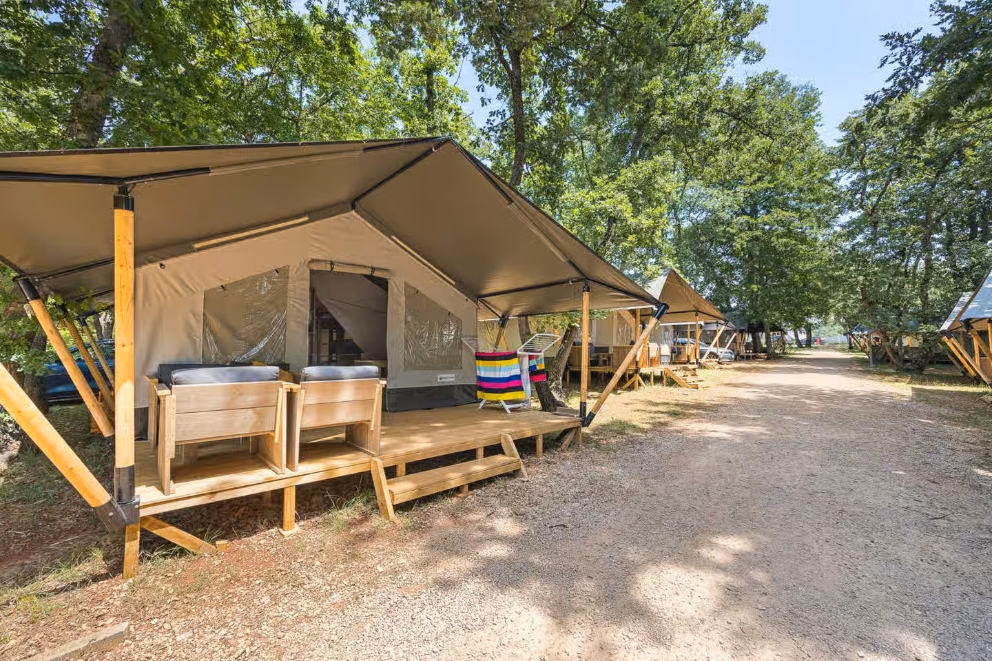 Rij safaritenten van Easyatent langs een grindpad op Camping Park Umag, met luifels op houten schoren en veranda's met loungestoelen onder de bomen