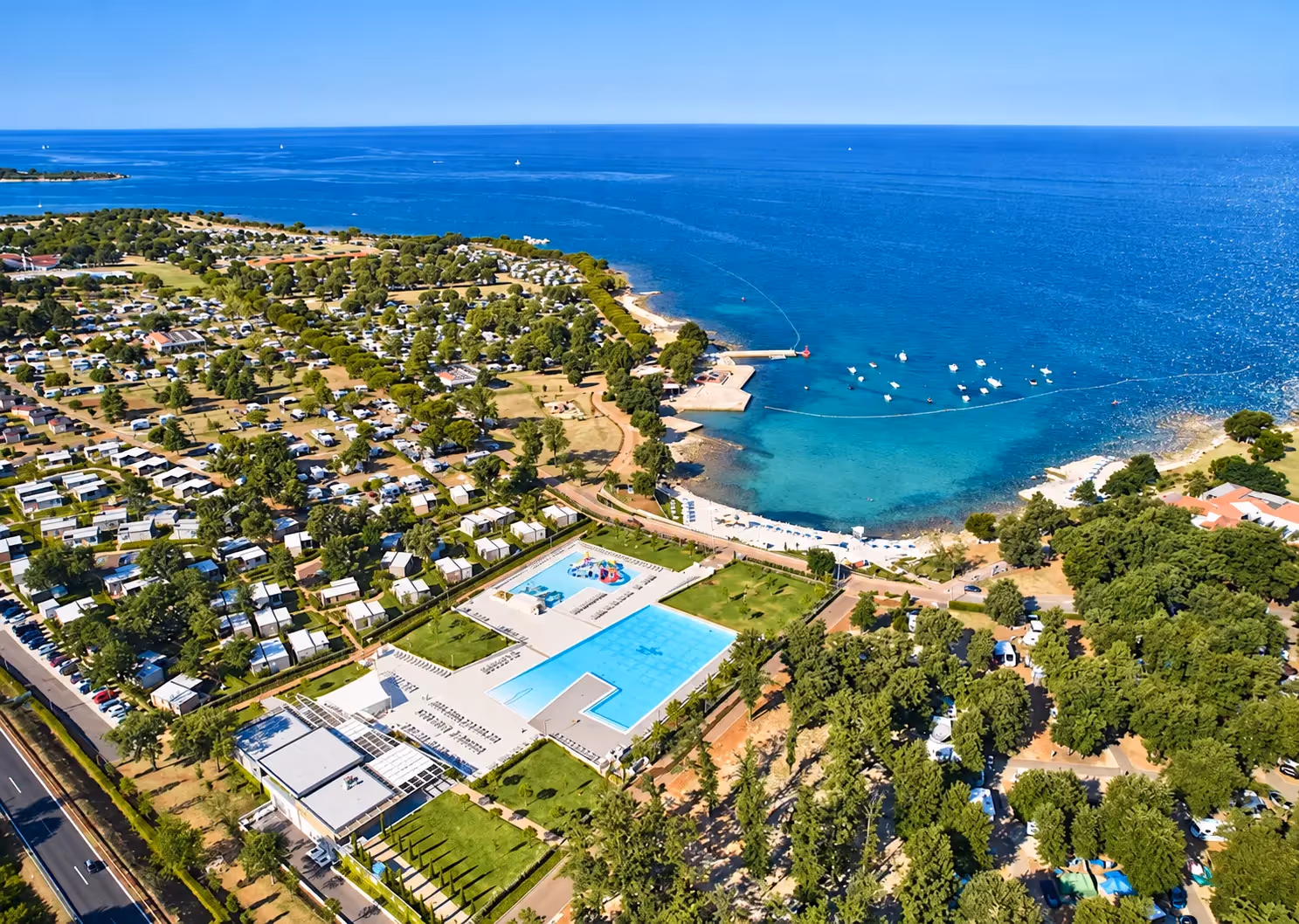 Luchtfoto van Camping Park Umag aan de Adriatische Zee, met het terrein vol staanplaatsen en mobilehomes tussen de bomen, de twee zwembaden en een steiger met bootjes in de baai