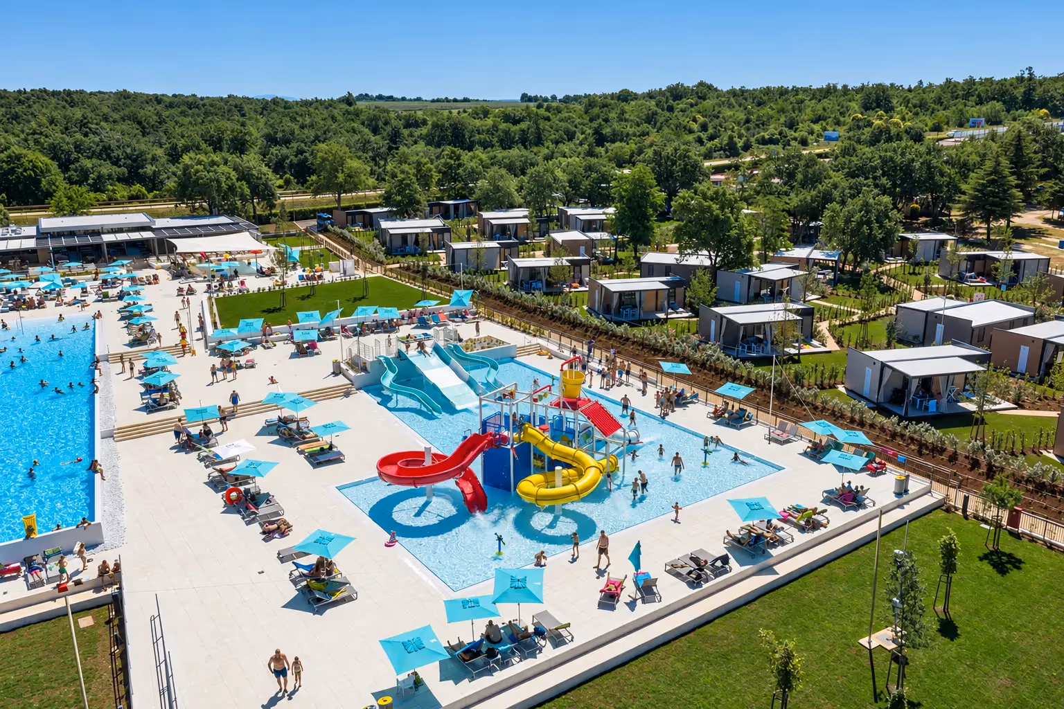 Luchtfoto van het waterpark van Camping Park Umag, met een kinderbad met een rode en een gele glijbaan, een groot zwembad met turquoise parasols eromheen en de rij mobilehomes erachter