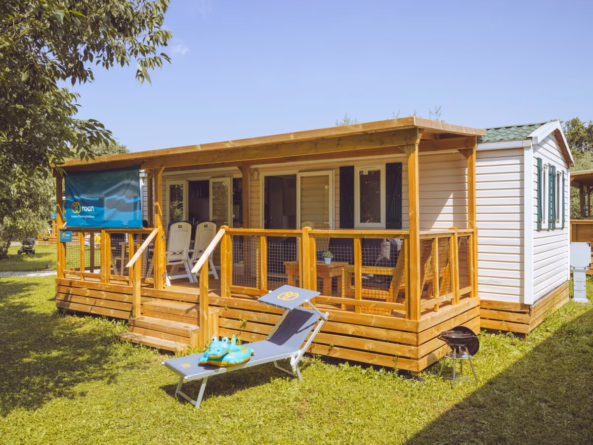 Stacaravan met een houten overkapping en veranda in het gras op Camping Park Umag, met een ligbed en slippers ervoor