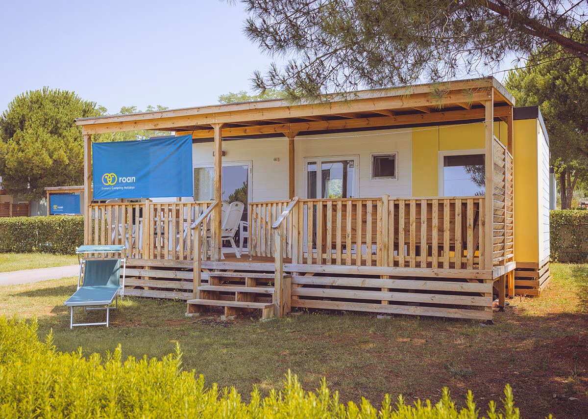 Geel-witte stacaravan met een houten overkapping en veranda op Camping Park Umag, met een ligbed in het gras ervoor